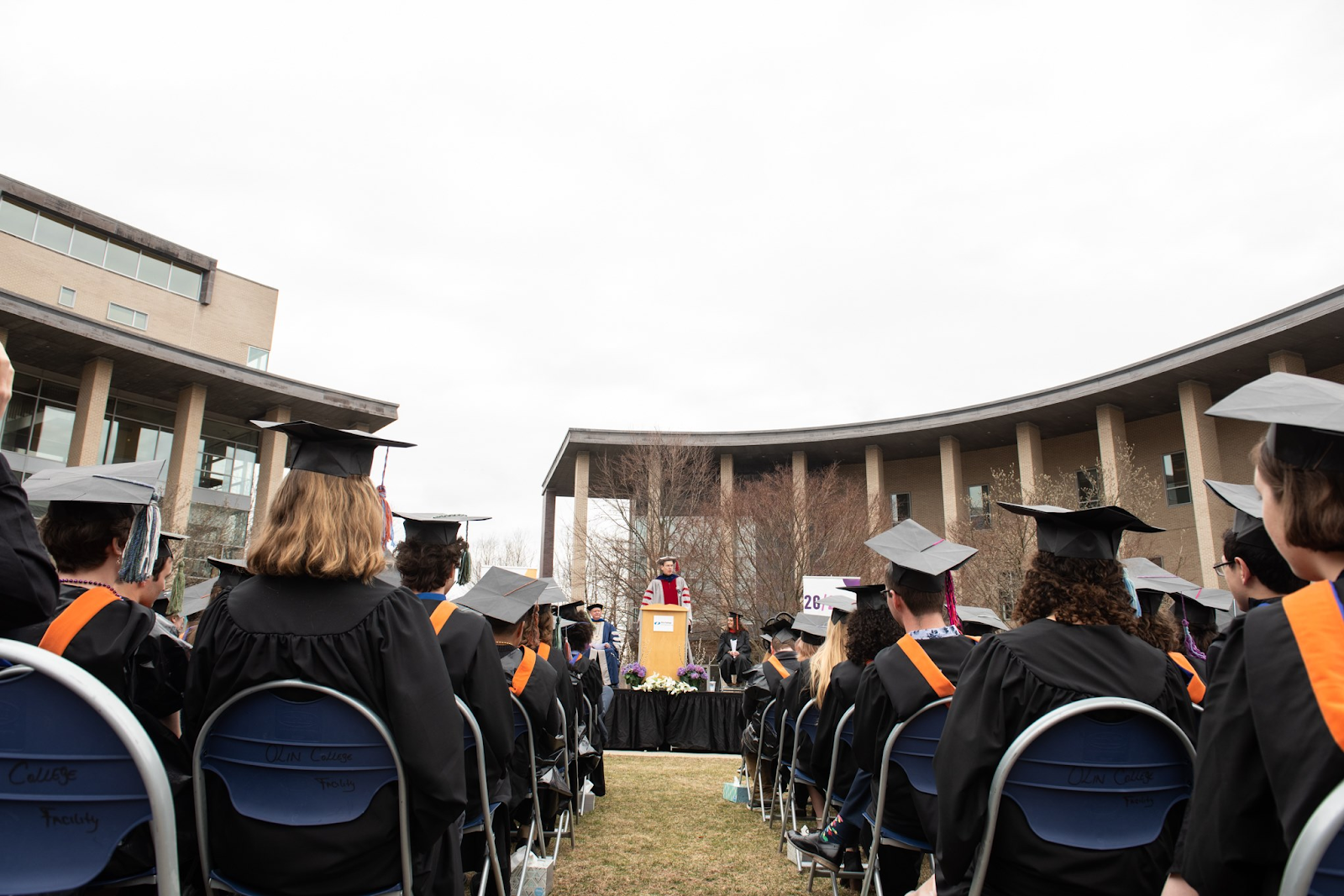 Students in graduation attire seated in rows in the Olin O, listening to a speaker.