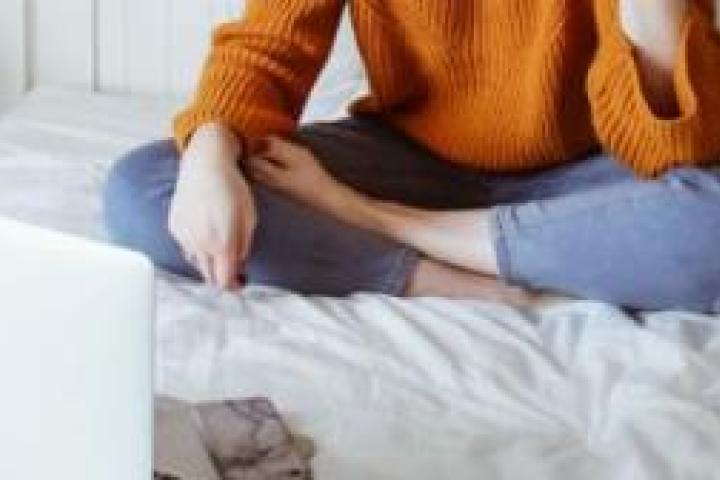 A closeup of a person sitting on a bed in front of a laptop.