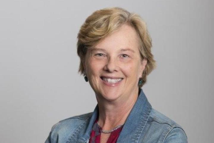 Woman with short blonde hair stands, smiling with arms crossed, wearing a jean jacket and red shirt.