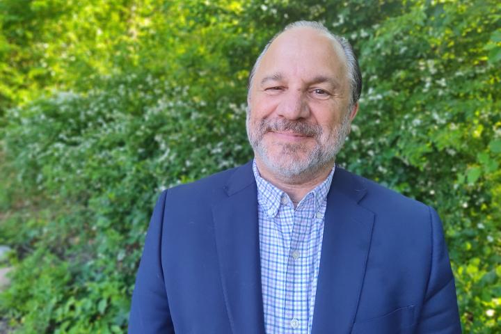 A photo of a man in a blue jacket standing in front of a green bush