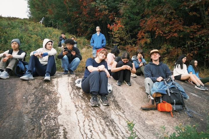 A large group of Olin College students sit on the side of a hill while camping at the White Mountains in NH.
