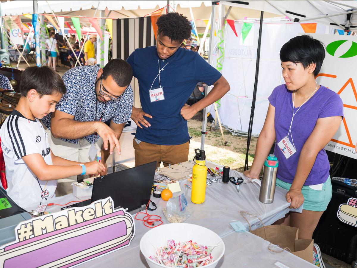 A man helps a young boy with a DIY coding project with a laptop and a tool made out of popsicle sticks with people looking on.