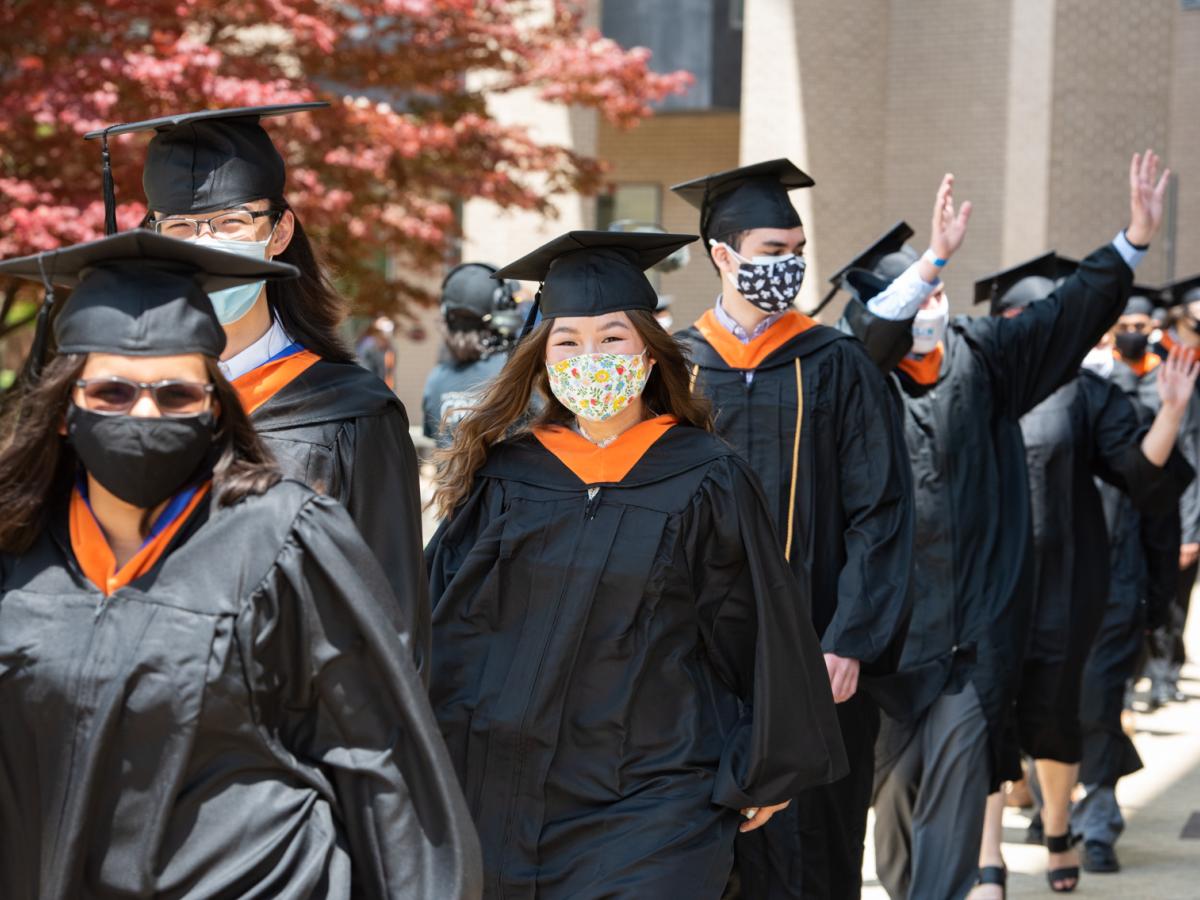 Members of the Olin College Class of 2021 wave to the community as they process to the Commencement Tent. A line of students in cap and gown wave and process down to the Olin Commencement Tent.
