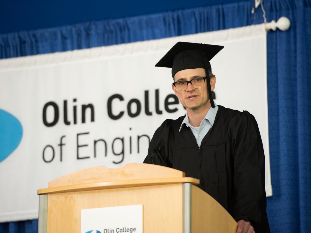 Tim Sauder, Associate Professor of Practice in Design, gives the Faculty Greeting on May 16, 2021 Tim Sauder, Associate Professor of Practice in Design, in a black cap and gown, stands at a podium with the Olin College logo behind him.