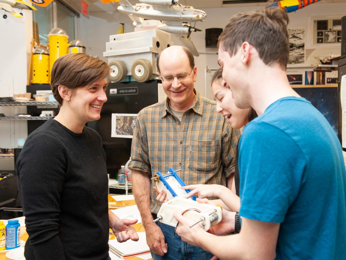 Professor Dave Barrett (in the center) with checkered shirt and glasses, and Professor Daniella Faas (on the left) in black shirt and close-cropped hair, talk with students in a machine lab in 2019.