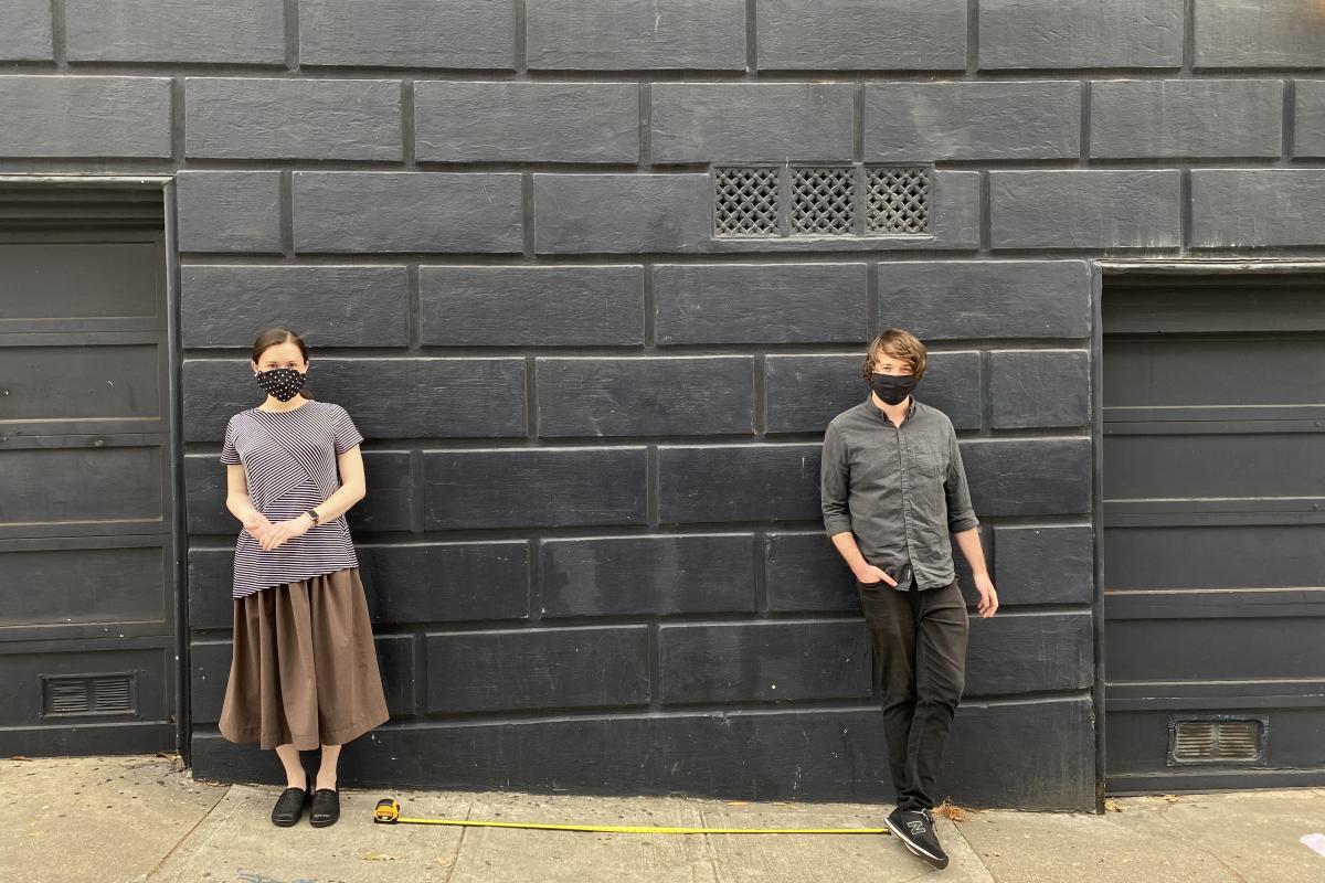 Brief's Andrea Huey (CTO) and Nick Hobbs '11 (CEO) pose (6 feet apart) recently. Two people stand against a grey, brick wall, approx. 6 feet apart.