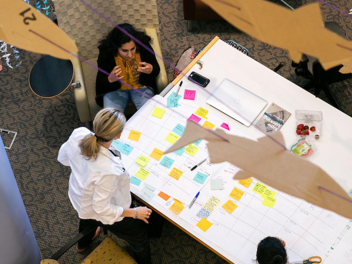 A scene from Sketch Model at Olin College. A birds-eye view of two people working on a white table, filled with sticky notes and a shark-like cut-out.