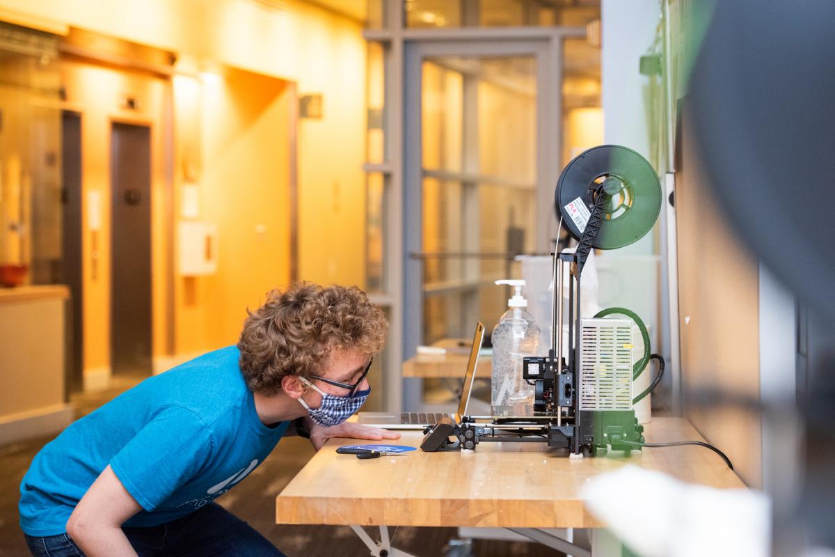 A student in blue shirt, mask and facial protection glasses bends over a table to look at a 3D printing machine.