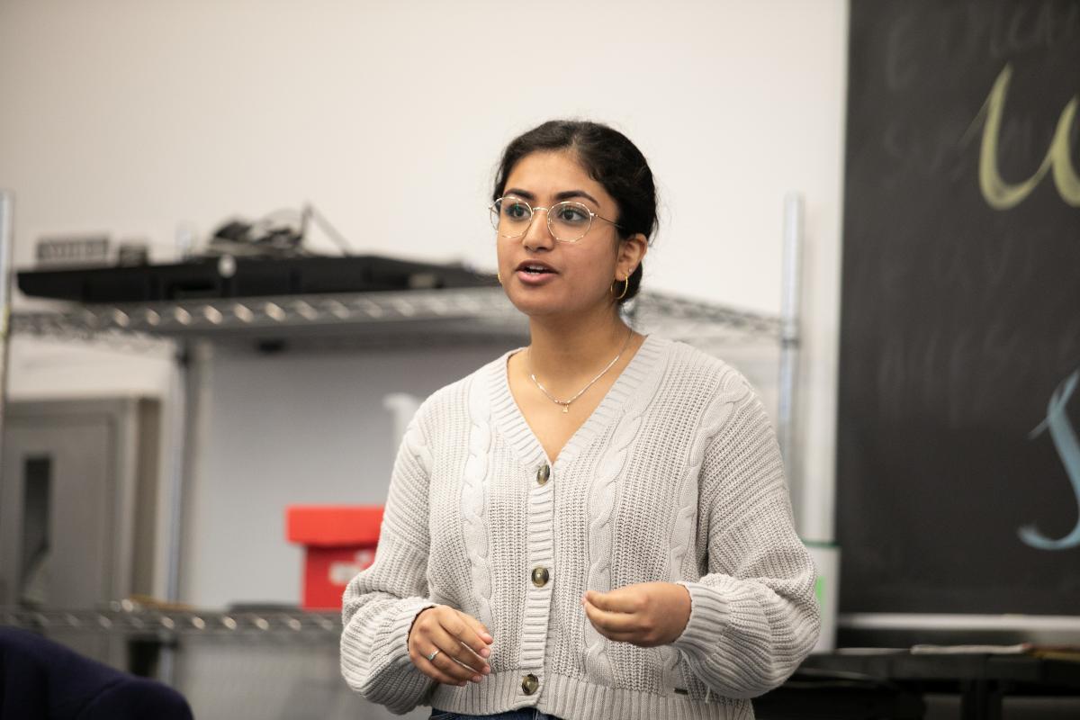 A student with black hair and light gray sweater and glasses talks in a classroom.