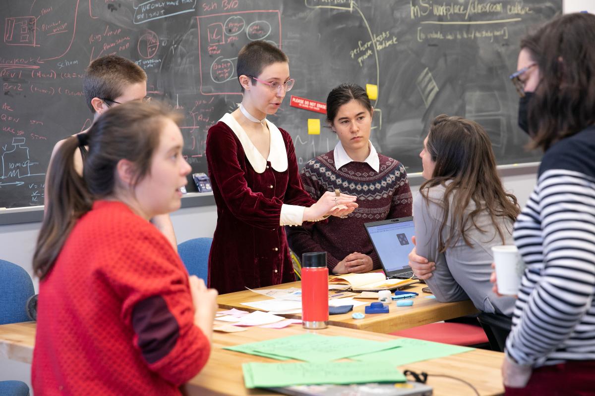 Multiple students collaborate with Professor Alessandra Ferzoco around a table in a classroom with a large blackboard in the background. Multiple students collaborate with a professor around a table in a classroom with a large blackboard in the background.