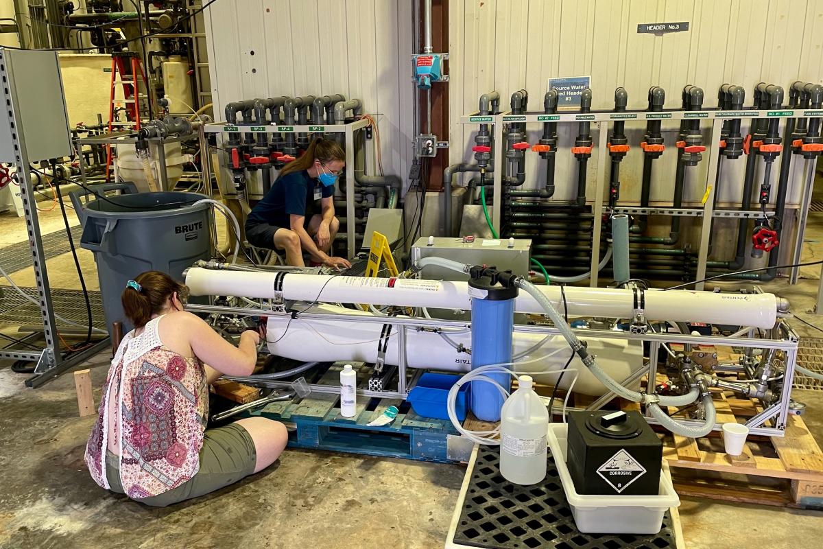 Emily W. Tow and Audrey Abraham '24 are taking in the concentrated waste stream from the Yuma Desalting Plant via their prototype desaltination system. Two people are sitting cross-legged working with their hands on two large white tubes.