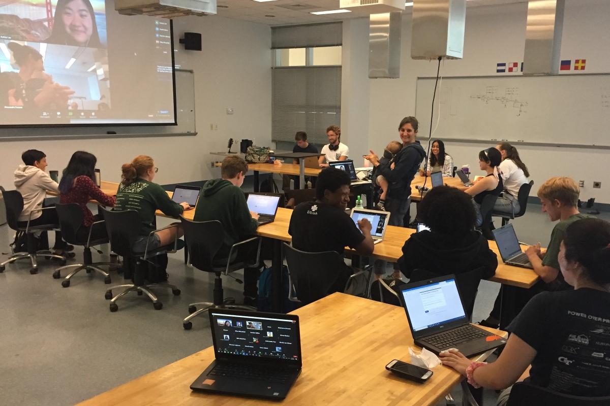 Olin Professor Alessandra Ferzoco is shown leading a scientific documentation workshop in 2021 at Olin.. A women holding a baby stands in the middle of a classroom with students around her working on laptops