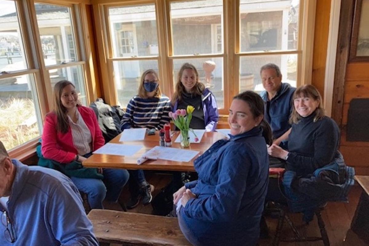 A group of seated people, look at the camera from around a wooden table.