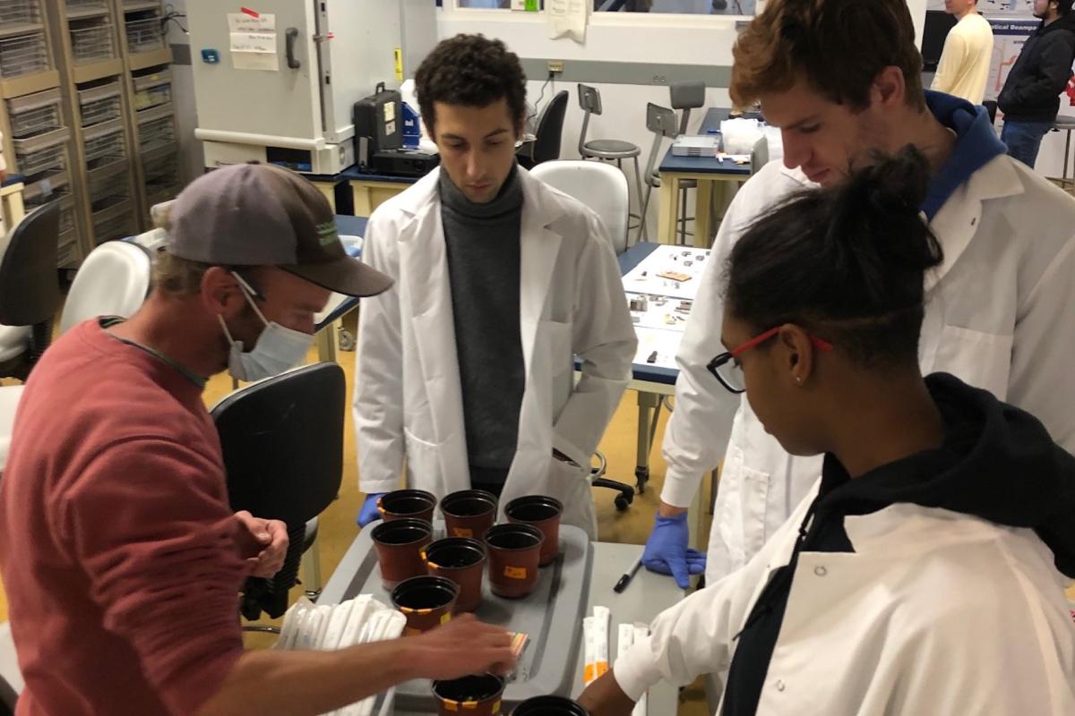 Three students in white lab coats watch as a worker from Black Earth Compost tests compost tea in little containers.