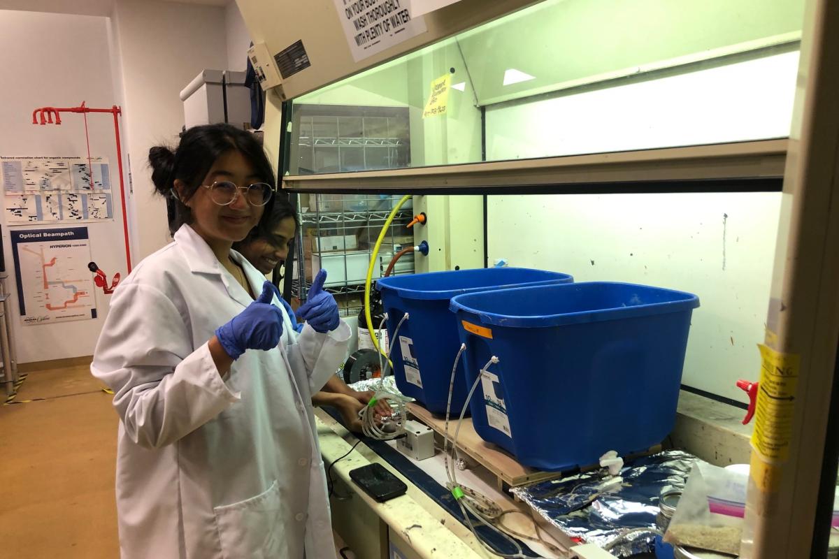 A student in a white lab coat and round glasses looks at camera and gives a thumbs up in a lab setting.