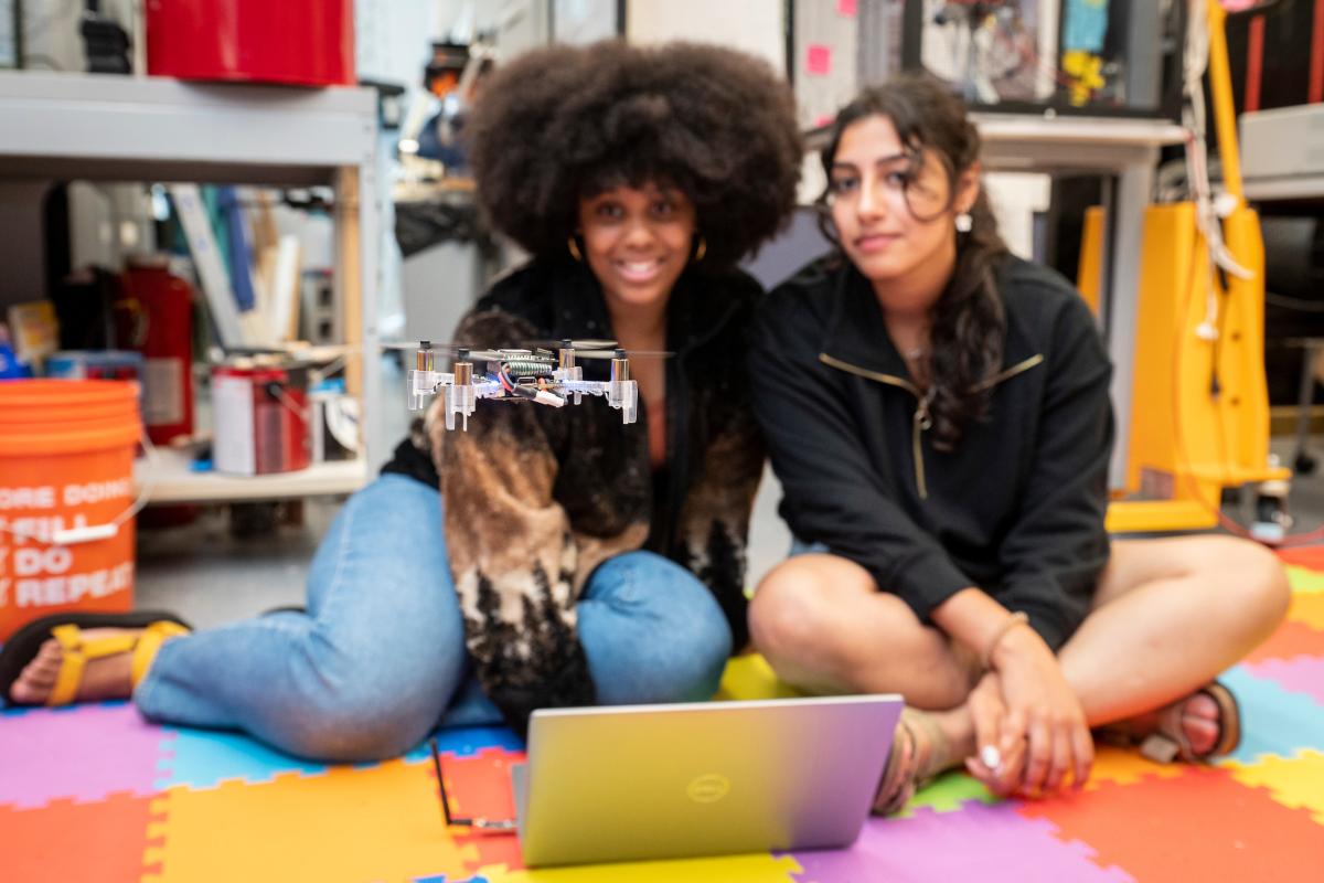 Two students sit on a colorful puzzle-piece mat in an Olin College classroom, while a small drone flies in front of them.