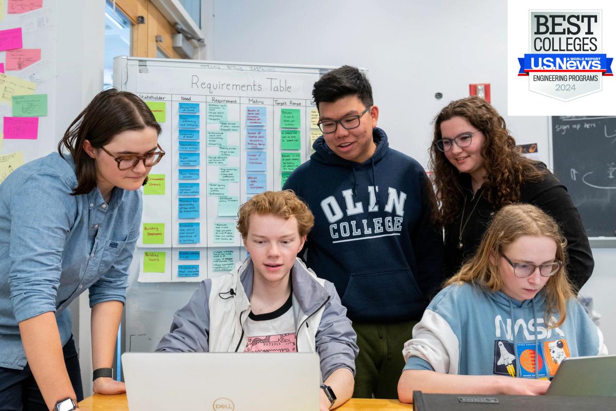 Five students work around two silver laptops in a classroom. Five students work around two silver laptops in a classroom.