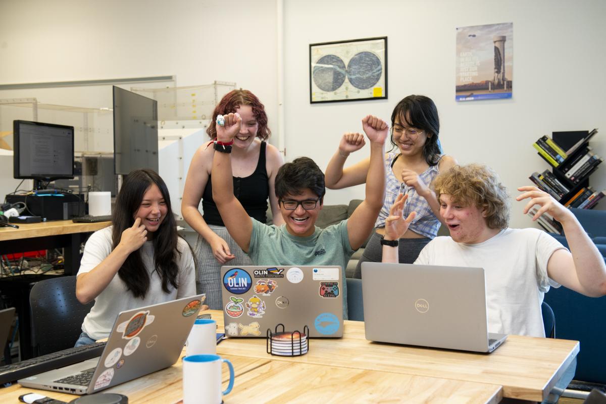 A group of five students in a collaborative workspace celebrate a success while working on laptops, smiling and cheering.