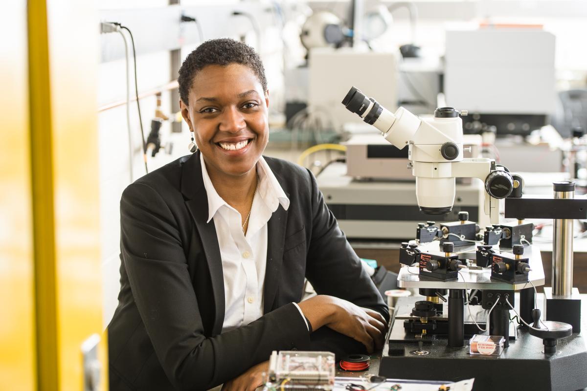 Dr. Shayla Sawyer Armand, Olin Provost, is pictured in a laboratory, sitting in front of a microscope and smiling at the camera. 