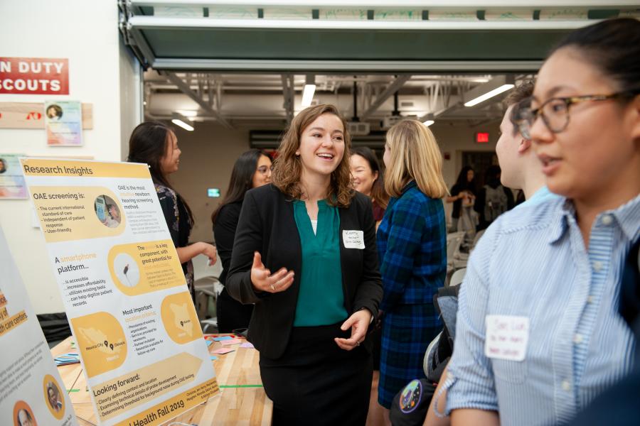 An Olin student talks with guests during an ADE poster presentation. A student in green blouse and black blazer talks with guests in front of a poster board.