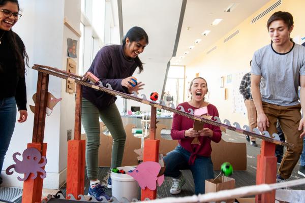 Four people laugh and smile while rolling a red ball down a slanted board with purple octopus cut-outs attached to the vertical pillars holding it up. 