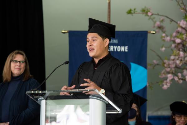 A young man stands at a podium wearing graduation regalia