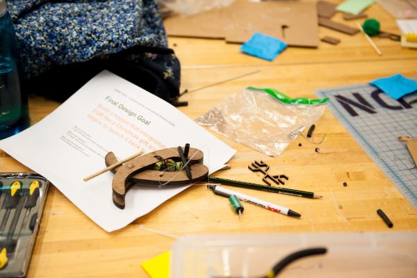An image of a table strewn with mechanical materials and a paper that reads "Final Design Goal."