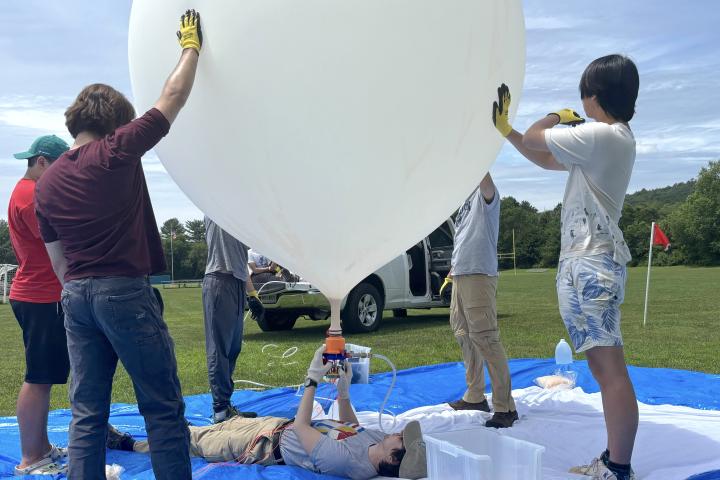 Olin students are holding up a large, white, high-altitude weather balloon as another team member lays flat on the ground inflating it. 