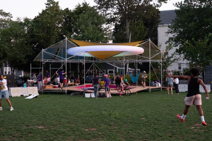 A platform with covers above it held up by metal poles. The white ring lights up at night. There are people throughout the photo talking and playing ball. 