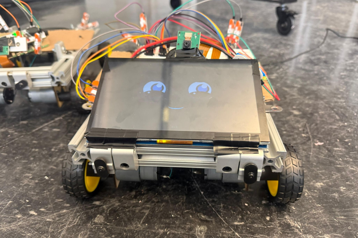 A small wheeled robot on a dark stone surface, with a tablet displaying a face consisting of two large eyes and a mouth