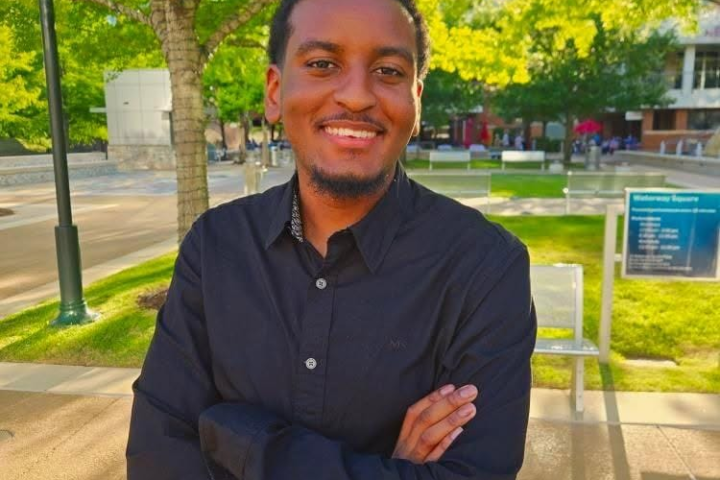 person with arms crossed stands outdoors smiling at camera