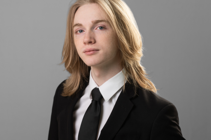 person in suit stands stoic in front of gray background