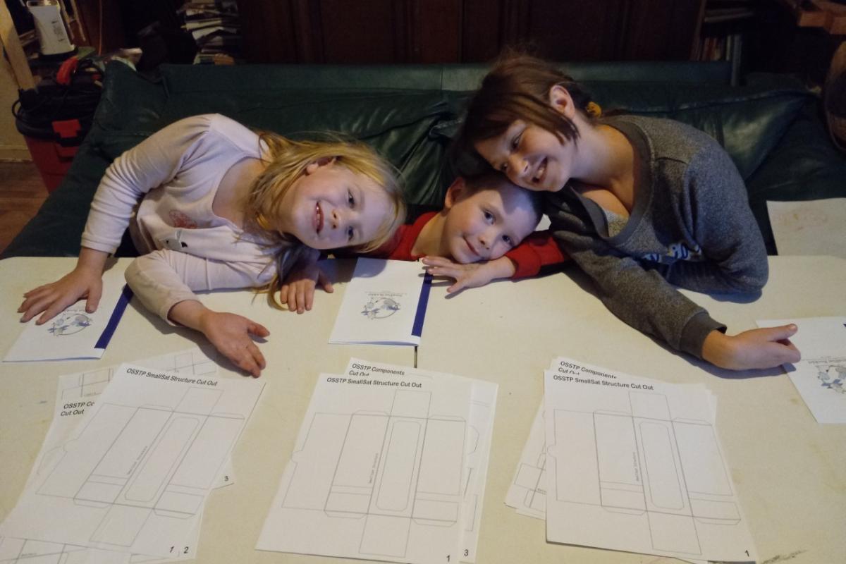 Three small children squat down in front of a small table with three piles of white papers spread out in front of them.