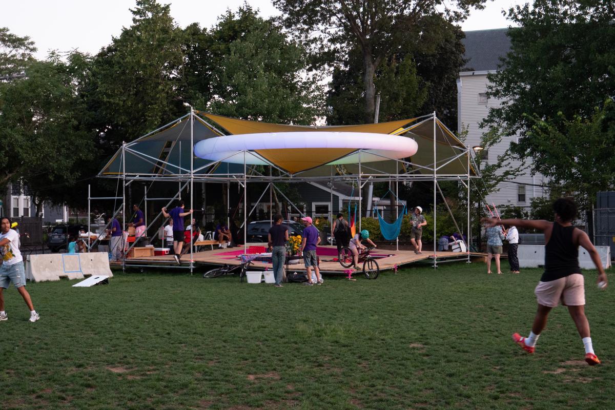 Shade is Social Justice structure. A platform with covers above it held up by metal poles. The white ring lights up at night. There are people throughout the photo talking and playing ball.
