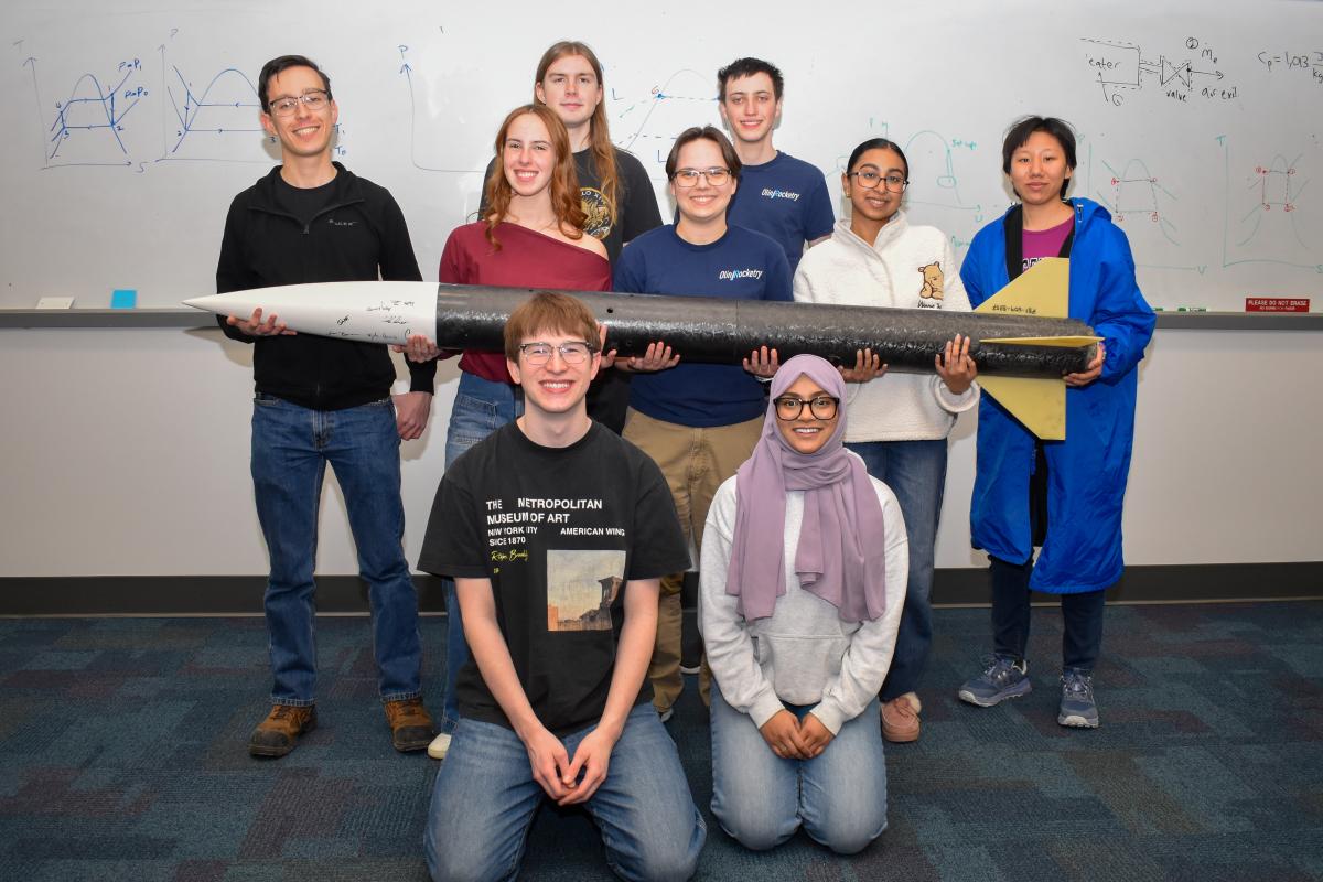 Olin Rocketry project team members pose for a group photo with a rocket in an Olin classroom.