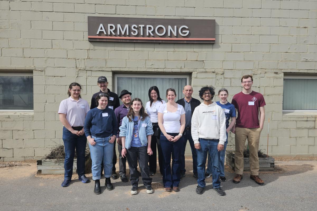 A group of staff and students stand in front of a building with a sign that reads "ARMSTRONG." 