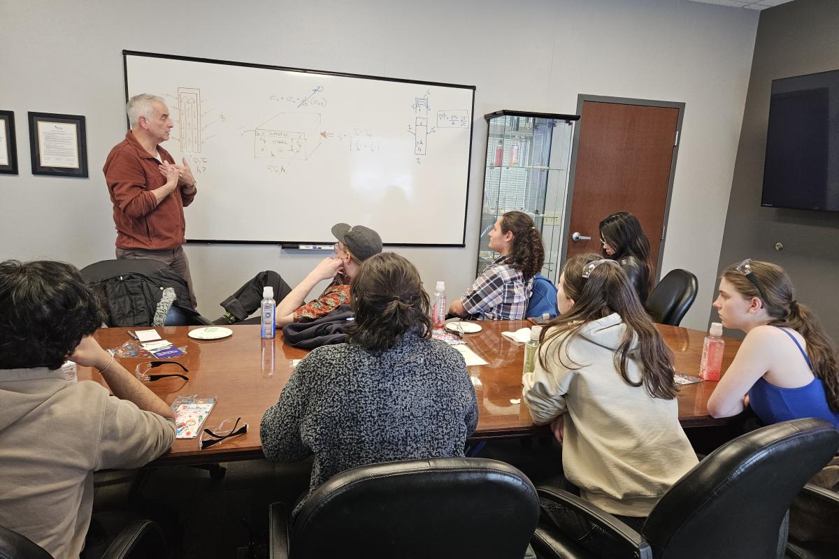 A man presents information on a white board to students and staff sitting around a brown conference table. 