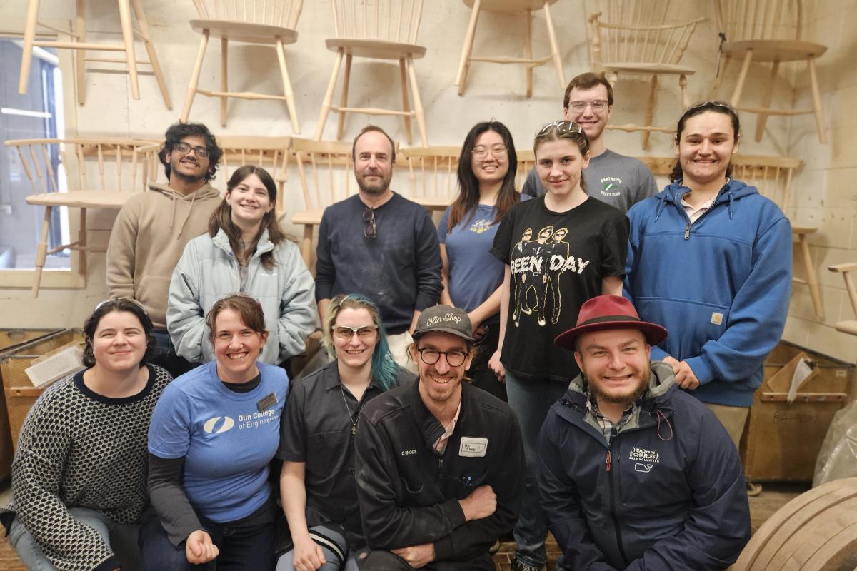 Students and staff pose in front of a pale wooden wall with stools.