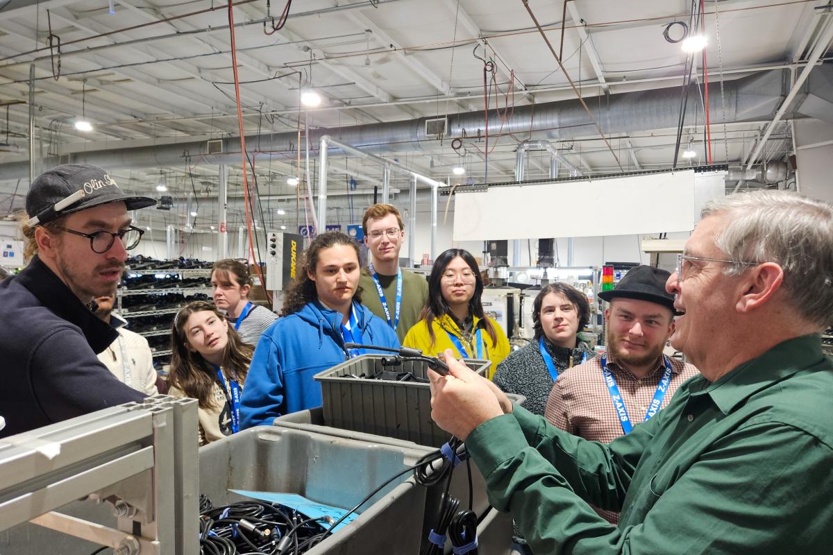 Students and Olin staff stand around a man demonstrating their process at Z-AXIS & Bear Power Supplies. 