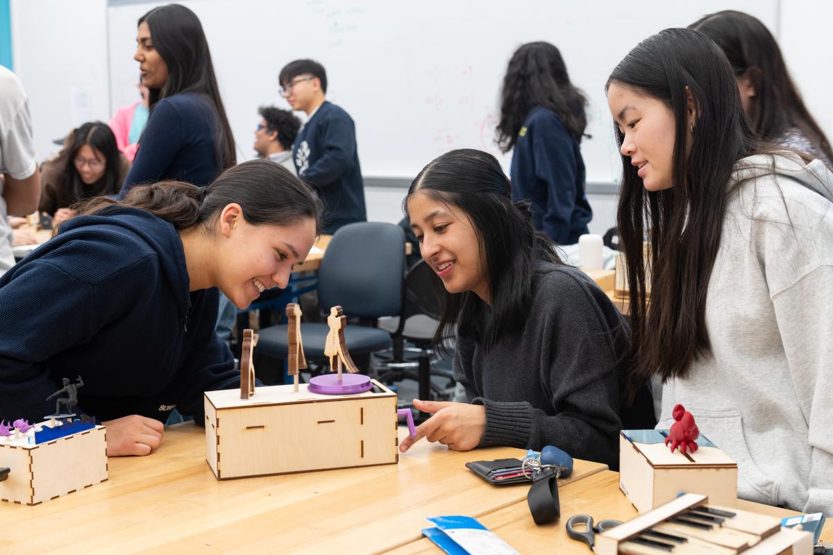 Three Olin College of Engineering students smile and laugh as they gather around a table to look at a final class project for Mechanical Prototyping. 