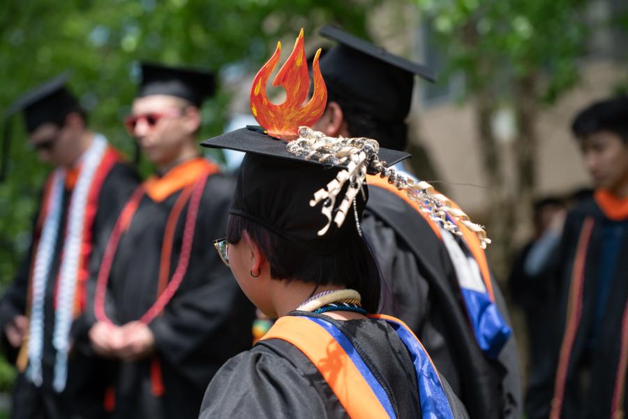 Commencement 2025, students in cap and gowns. Students fire decorated hat.
