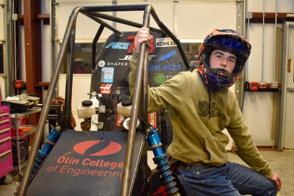 An Olin Baja project team student sits on the car wearing a helmet and poses for the photo.