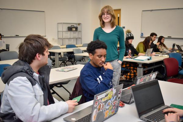Three Olin OAT project team members, two sitting and one standing, work together on a project with laptops.