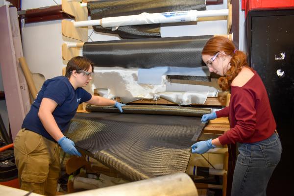 Two Olin Rocketry project team members work together to cut a length of grey fabric from a roll.