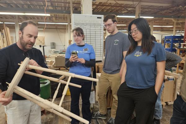 Students and a staff member stand around a O&G Studio employee showing them a pale wooden stool. 